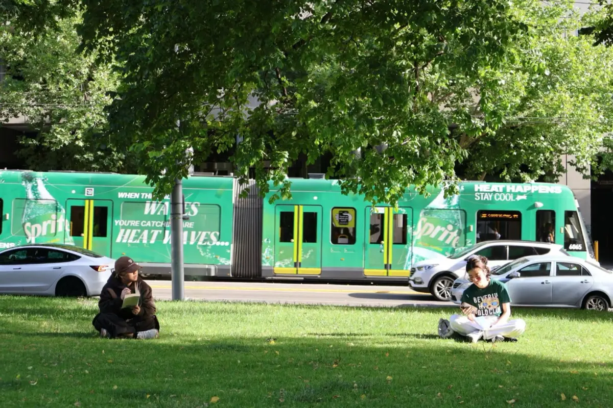 A green Melbourne tram passing a park lawn where people sit in the shade